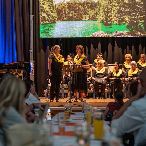 Ein Chor tritt auf der Bühne mit einer landschaftlichen Kulisse eines Sees auf. Zwei Frauen stehen vor einem Klavier und einem Bildschirm, während andere hinter ihnen sitzen. Das Publikum beobachtet von Tischen mit Getränken aus.