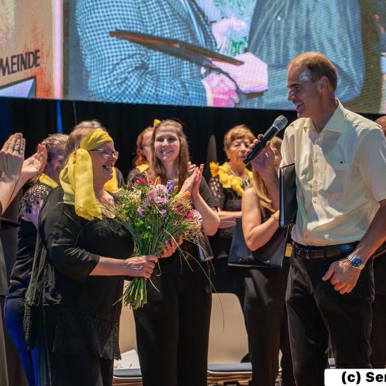 Eine Frau hält Blumen und klatscht, während ein Mann in ein Mikrofon spricht bei einer Veranstaltung. Andere Teilnehmer klatschen und lächeln. Eine große Leinwand zeigt einen Mann mit einer Gitarre.