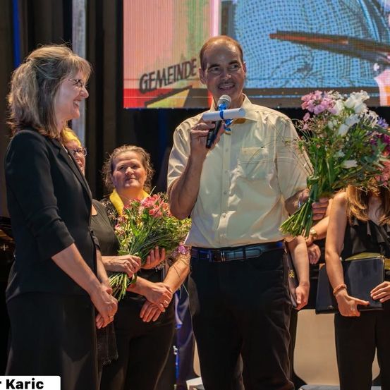Ein Mann hält ein Mikrofon und scheint eine Rede zu halten, während er neben einer Frau steht, die Blumen hält. Dahinter hält eine weitere Frau einen Blumenstrauß, und es gibt einen Bildschirm im Hintergrund.