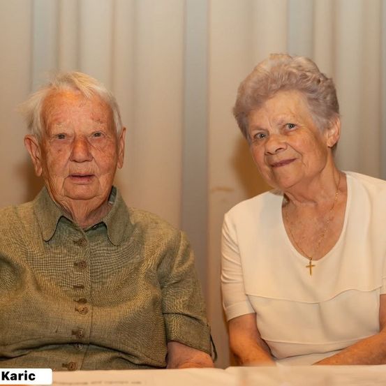Ein älteres Paar mit weißen Haaren sitzt nebeneinander am Tisch. Der Mann trägt ein grünes Hemd und die Frau ein weißes Oberteil mit Kreuzkette. Hinter ihnen ist ein weißer Vorhang.
