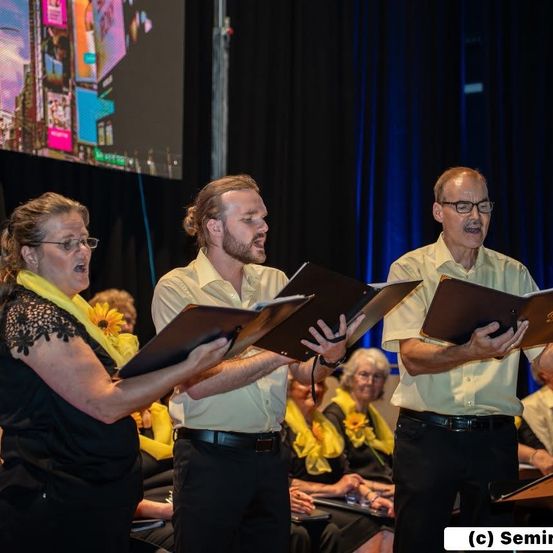 Drei Personen, zwei Männer und eine Frau, stehen vor einer Menge, halten Notenbücher in den Händen und singen. Hinter ihnen zeigt eine große Leinwand verschiedene Bilder. Das Publikum, das im Hintergrund sitzt, beobachtet aufmerksam.
