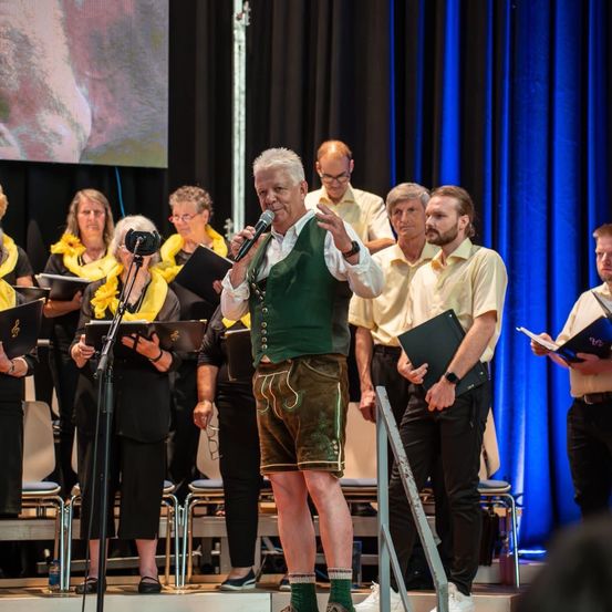 Ein Mann steht auf der Bühne und singt in ein Mikrofon. Hinter ihm halten mehrere Menschen Bücher und singen. Blaue Vorhänge sind im Hintergrund.