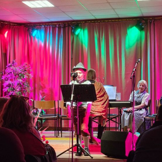 Ein Künstler in einem Hut singt auf der Bühne mit einer Pianistin. Die Bühne ist mit roten und grünen Lichtern beleuchtet. Ein Publikum sitzt vor der Bühne.
