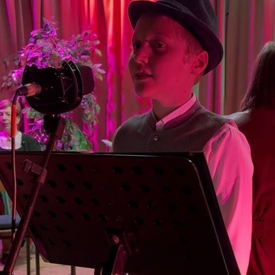 Ein junger Junge steht vor einem Mikrofon und singt möglicherweise bei einer Veranstaltung. Er trägt einen schwarzen Hut und ein rosa Hemd. Hinter ihm steht eine Pflanze mit lila Blüten und eine sitzende Frau.