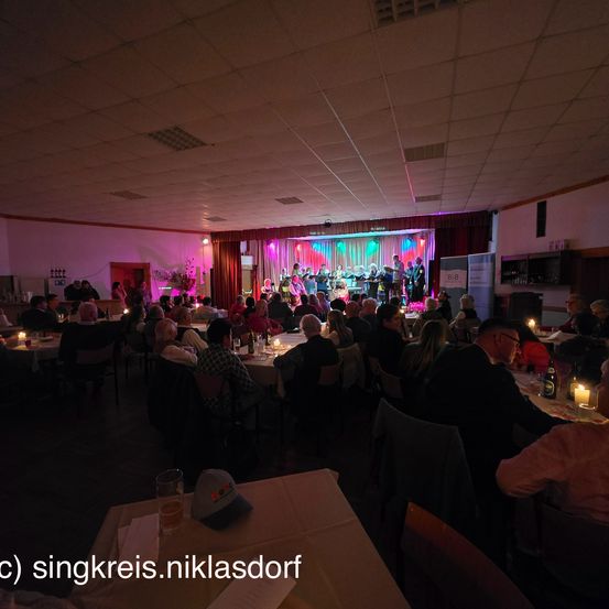 Eine nächtliche Indoor-Veranstaltung zeigt eine Band auf der Bühne, mit Tischen und Stühlen für das Publikum. Die Bühne ist mit mehrfarbigen Lichtern beleuchtet.