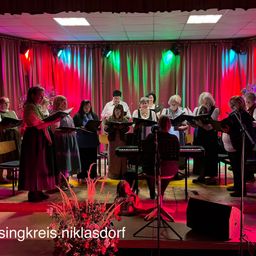 Ein Chor von Frauen steht auf der Bühne, hält ihre Notenblätter in den Händen, mit Keyboards und Mikrofonen. Sie sind von farbigen Bühnenlichtern beleuchtet, und unten befindet sich ein Wasserzeichen.