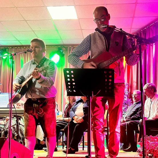 Zwei Männer spielen mit Gitarren auf der Bühne unter pinken und grünen Lichtern, mit einem Publikum vor ihnen sitzend.