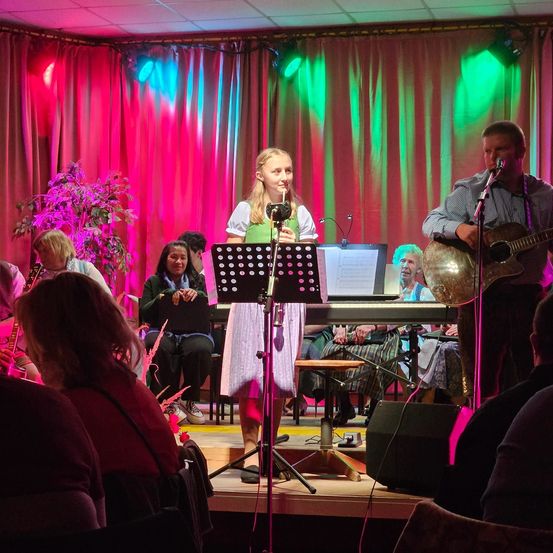 Eine Bühnenaufführung mit einer singenden Frau, einem Gitarre spielenden Mann und anderen Instrumentenspielern. Ein roter Vorhang und Scheinwerfer im Hintergrund.