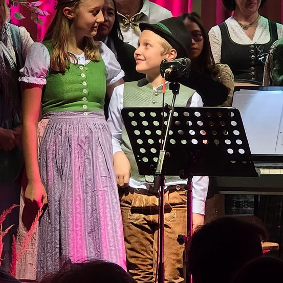 Ein Mädchen und ein Junge führen mit einem Mikrofon vor ihnen auf der Bühne auf. Sie tragen traditionelle Kleidung. Hinter ihnen stehen andere Leute, von denen einer Klavier spielt.