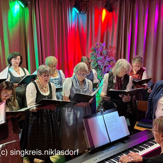 Eine Gruppe älterer Frauen singt im Chor, gekleidet in traditioneller Kleidung. Sie halten Notenblätter in den Händen und schauen darauf. Ein Klavier steht vor ihnen, und bunte Lichter beleuchten die Bühne.