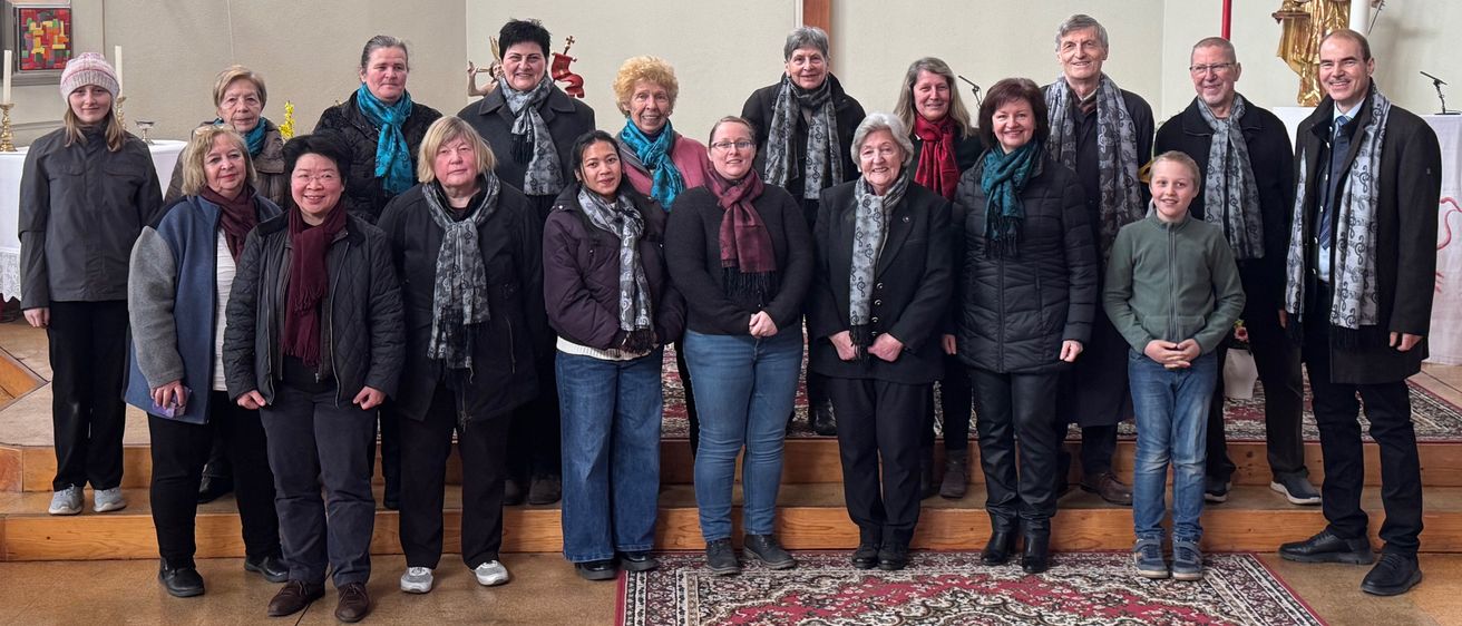 Eine Gruppe von Frauen in Winterkleidung, lächelnd, steht vor einer Kircheninneneinrichtung, mit einer Statue von Jesus und einem Kreuz im Hintergrund.