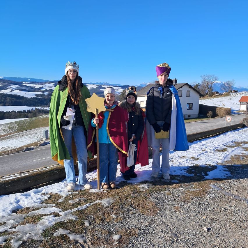 Vier Menschen in Kostümen, wahrscheinlich für ein Theaterstück oder eine Veranstaltung, stehen in einem verschneiten Bereich mit Bergen im Hintergrund.