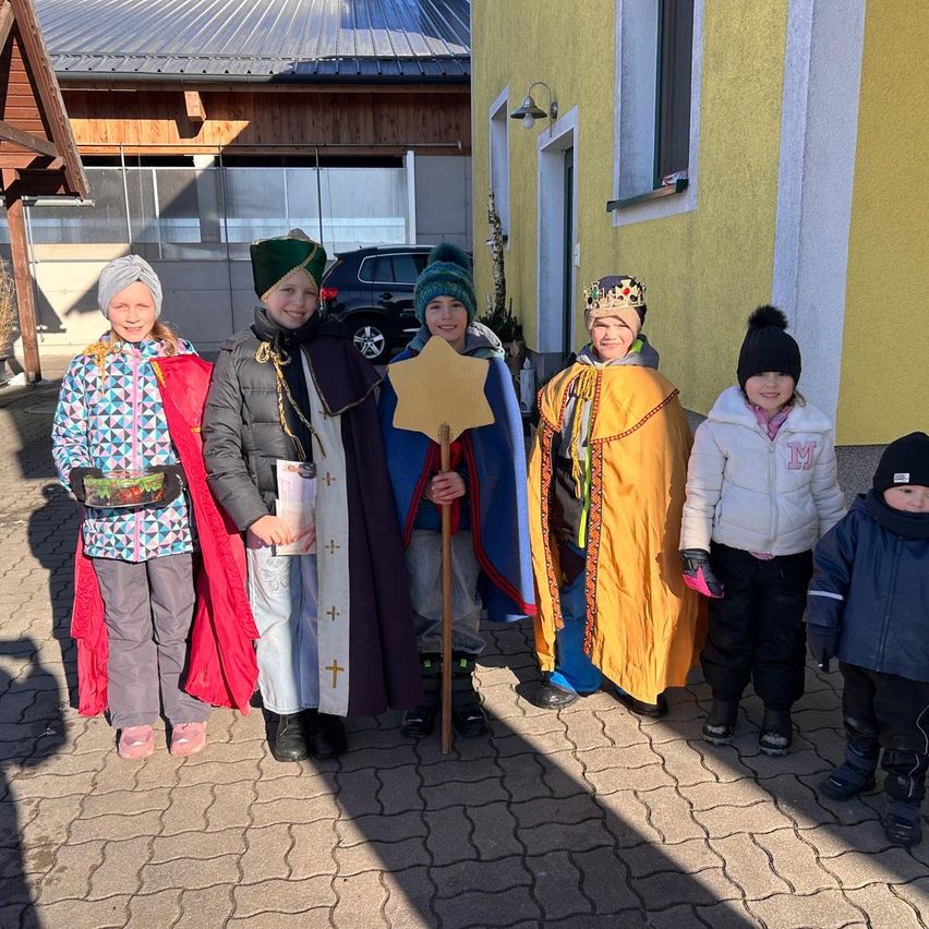 Eine Gruppe von Kindern in bunten Kostümen posiert für ein Foto vor einem gelben Gebäude. Sie halten einen Stern und ein Buch, und das Gebäude hat ein Fenster und eine Lampe.