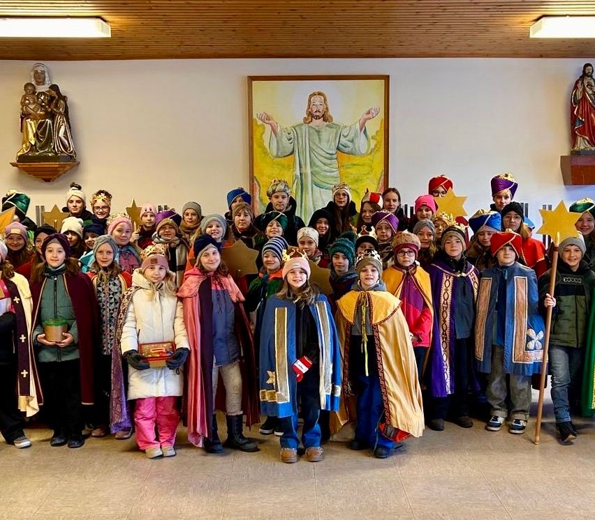 Eine Gruppe von Kindern in farbenfrohen Kostümen posiert für ein Foto in einem Raum mit einem religiösen Gemälde im Hintergrund.