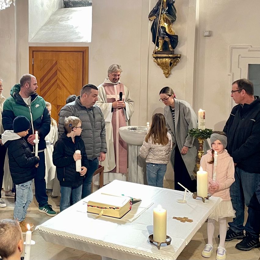 Eine Gruppe von Menschen steht um einen Tisch in einer Kirche. Ein Priester hält ein Mikrofon und spricht. Sie halten Kerzen. Eine Statue ist an der Wand montiert.