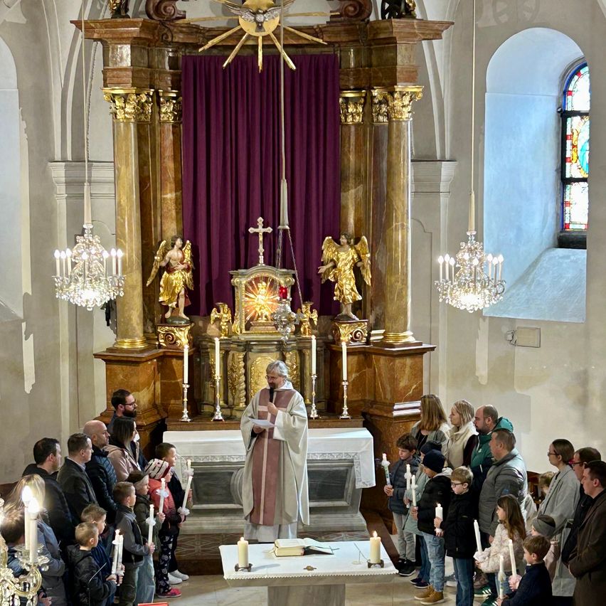 Ein Priester steht vor einem Altar in einer Kirche und liest aus einem Buch. Menschen, einschließlich Kinder, stehen und halten Kerzen. Der Altar hat ein Kreuz, Kerzen und Statuen.