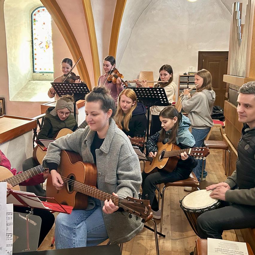 In einer Kirche spielt eine Gruppe junger Musiker Instrumente. Geigen, Flöten und Gitarren sind sichtbar. Eine Frau in einer grauen Jacke spielt die Gitarre. Eine andere Frau spielt eine Trommel. Ein Mann in einer Jacke steht in der Nähe.