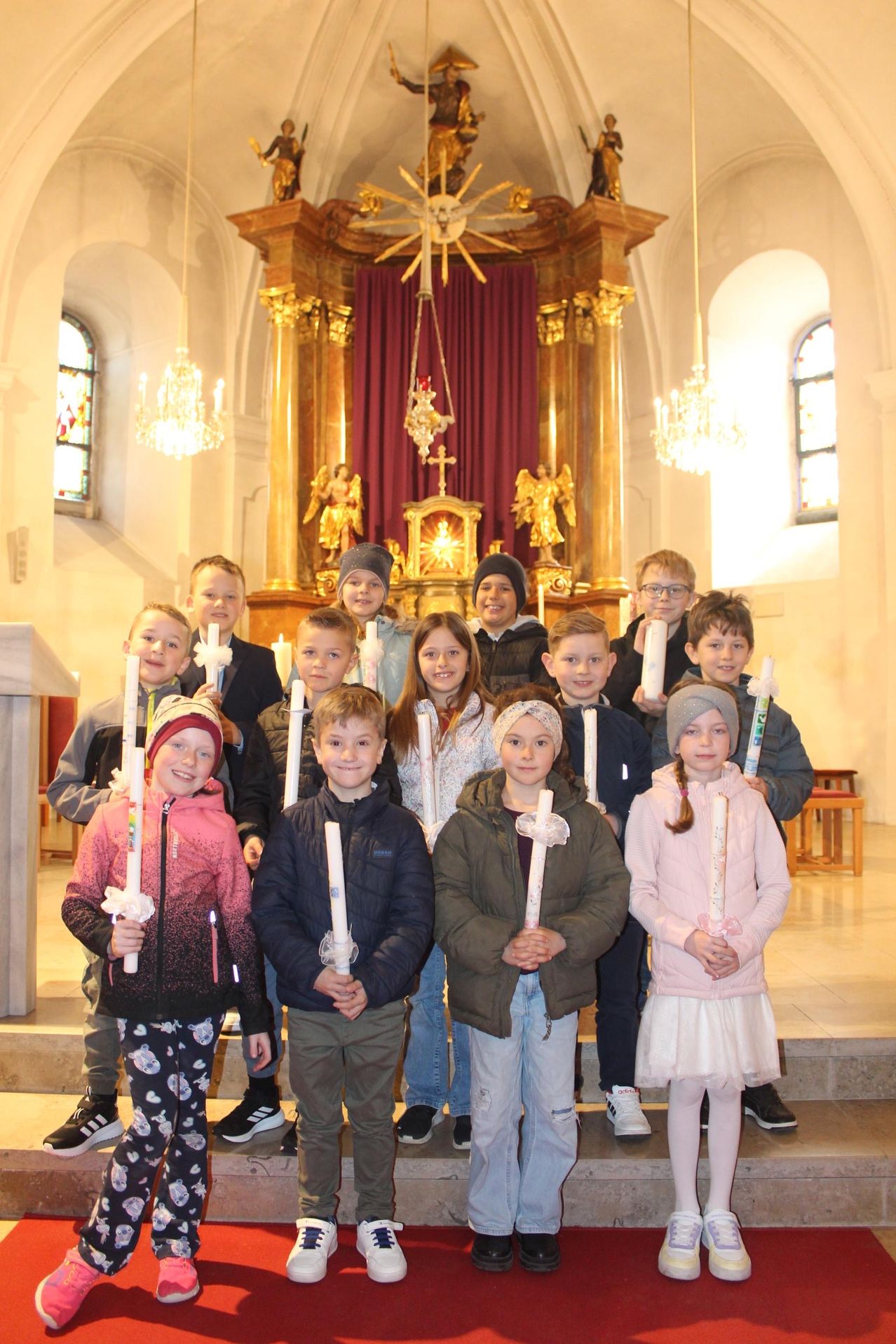 Kinder in Winterkleidung stehen in einer Kirche und halten Kerzen vor einem Altar.