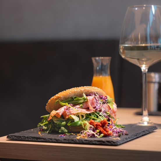 Ein Sandwich mit Fleisch, Gemüse und Sauce steht auf einem schwarzen Schieferteller mit einem Glas Wein auf einem Holztisch.