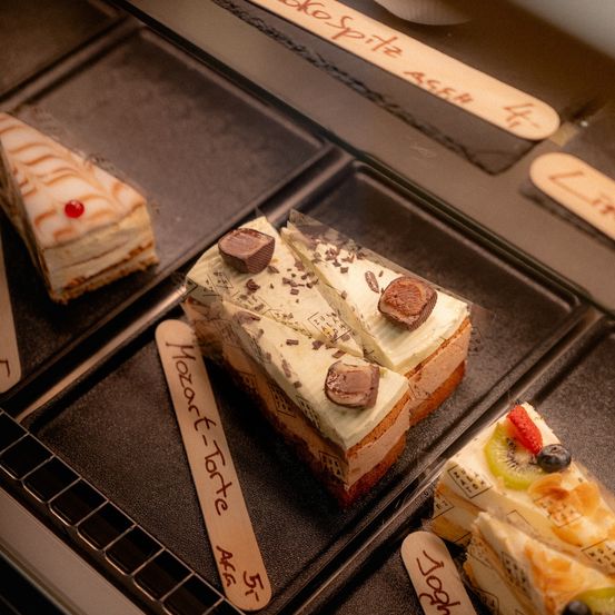 Eine Vitrine mit verschiedenen Desserts, darunter ein schokoladenüberzogener Kuchen, ein Stück Obsttarte und ein weiterer Kuchen mit einer Kirsche obenauf. Holzstäbchen dienen zur Identifizierung.