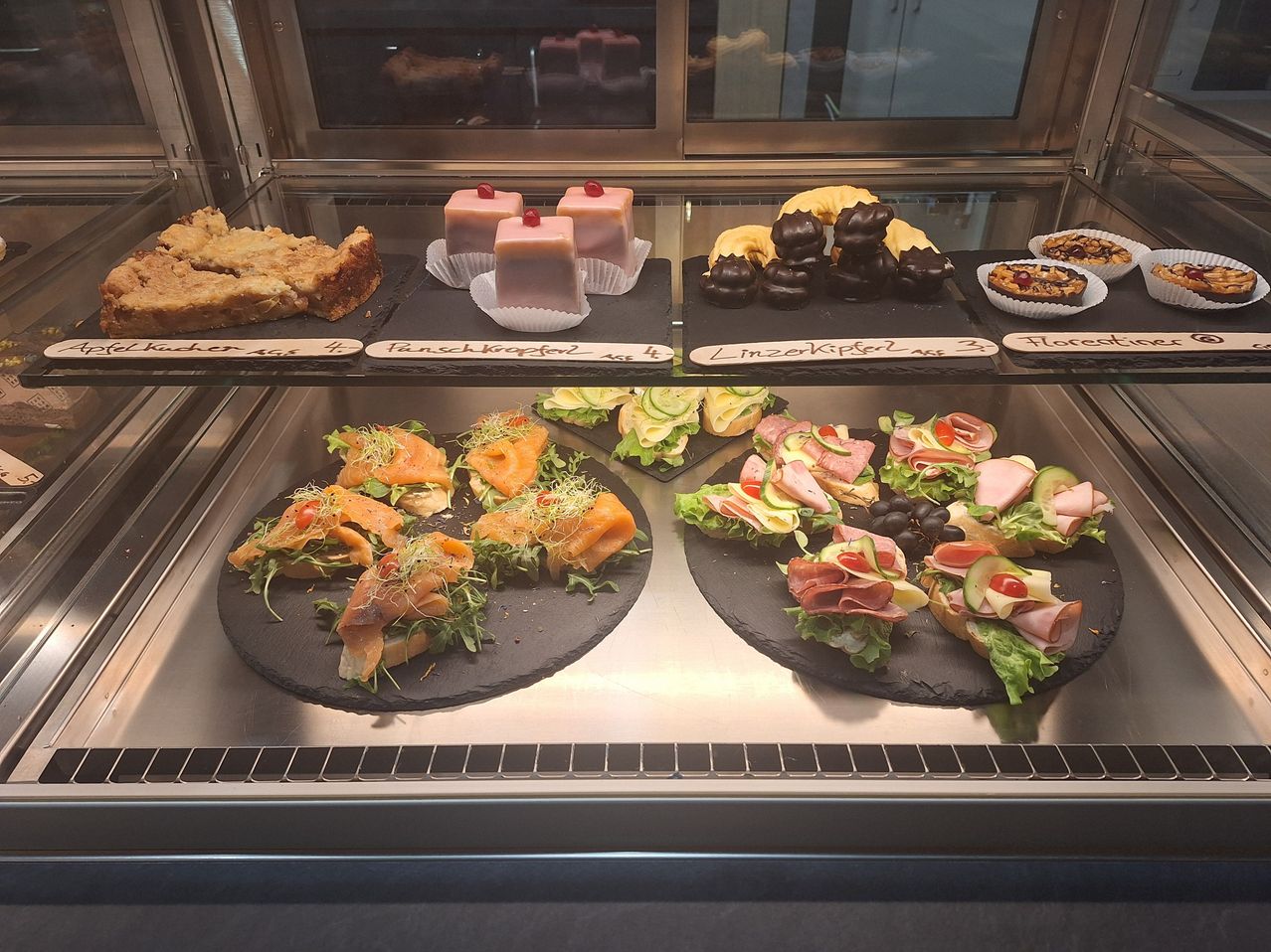 Eine Vitrine mit verschiedenen Gebäck und Sandwiches. Mehrere Tabletts halten verschiedene Arten von Sandwiches und Desserts, alle ordentlich angeordnet. Zu den Desserts gehören schokoladenüberzogene Gebäck.