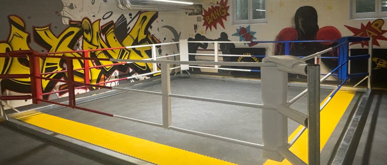Ein Indoor-Boxring mit Graffiti-Wänden, Metallgittern und einem weißen Boden. An der Wand sind die Wörter 'BANG' und 'BAM' gemalt.