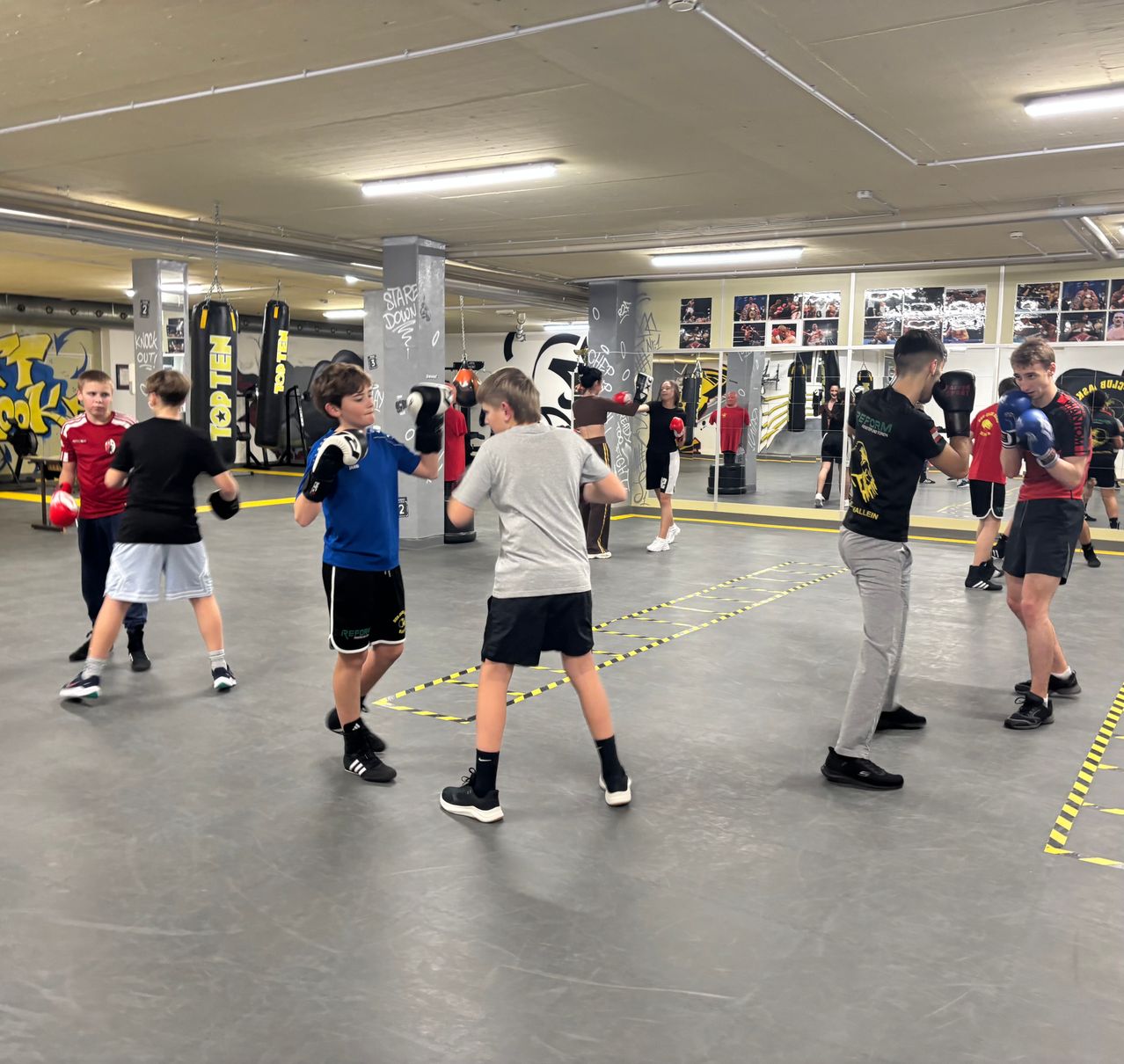 Eine Gruppe von Jungen und jungen Männern, die Boxhandschuhe tragen, übt Boxen in einem Fitnessstudio. Sie stehen paarweise gegenüberstehend.