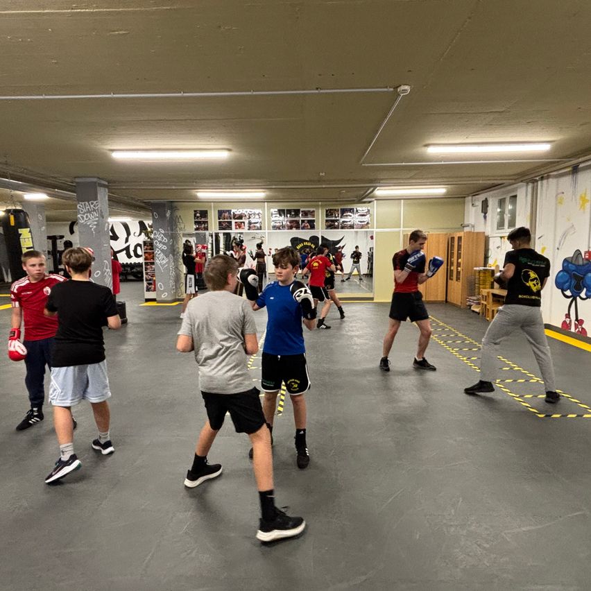 Mehrere junge Männer boxen in einem Fitnessstudio. Einige sind zu zweit, andere allein. Das Fitnessstudio hat eine mit Graffiti verzierte Wand, eine Decke mit Lichtern und einen Spiegel.
