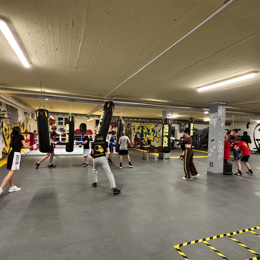 Eine Gruppe von Menschen trainiert Boxen in einem Fitnessstudio mit Graffiti an den Wänden. Sie schlagen auf schwere Boxsäcke ein.