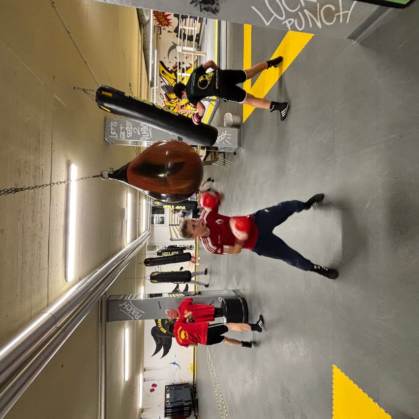 Ein Fitnessstudio mit mehreren Personen, die Boxaktivitäten durchführen. Eine Person in einem roten Shirt und Handschuhen schlägt auf einen Boxsack. Eine andere Person trägt ein schwarzes Outfit. Die Wände sind mit Graffiti und einer gelben Linie verziert.