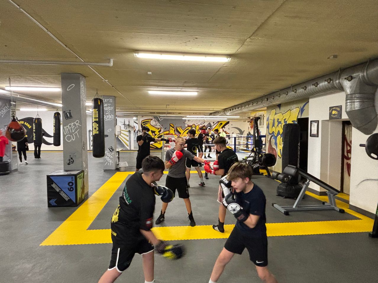 Ein Boxstudio mit Graffiti-Wänden hat mehrere Menschen, die Boxen üben, wobei einige Schlagsäcke benutzen. Einige Leute tragen Handschuhe und Kopfschutz.