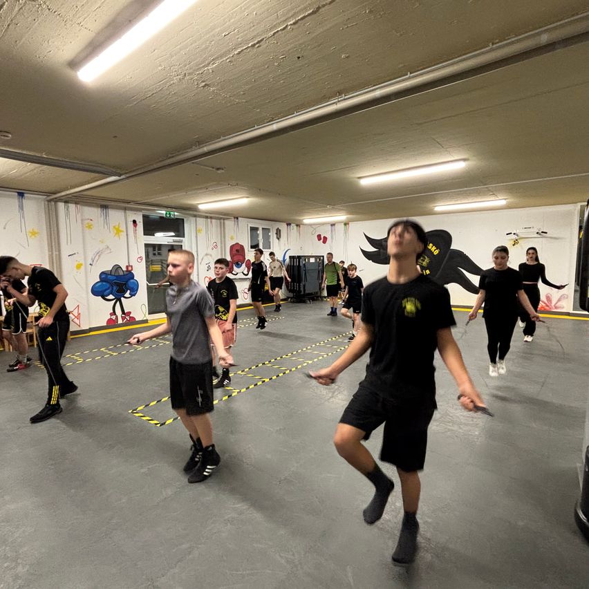 In einem Indoor-Fitnessstudio übt eine Gruppe von Menschen das Seilspringen, einige tragen schwarze und graue Uniformen. Der Boden hat farbenfrohe Graffiti und gelbe Linien. Eine Person in einem schwarzen Shirt schaut nach oben.