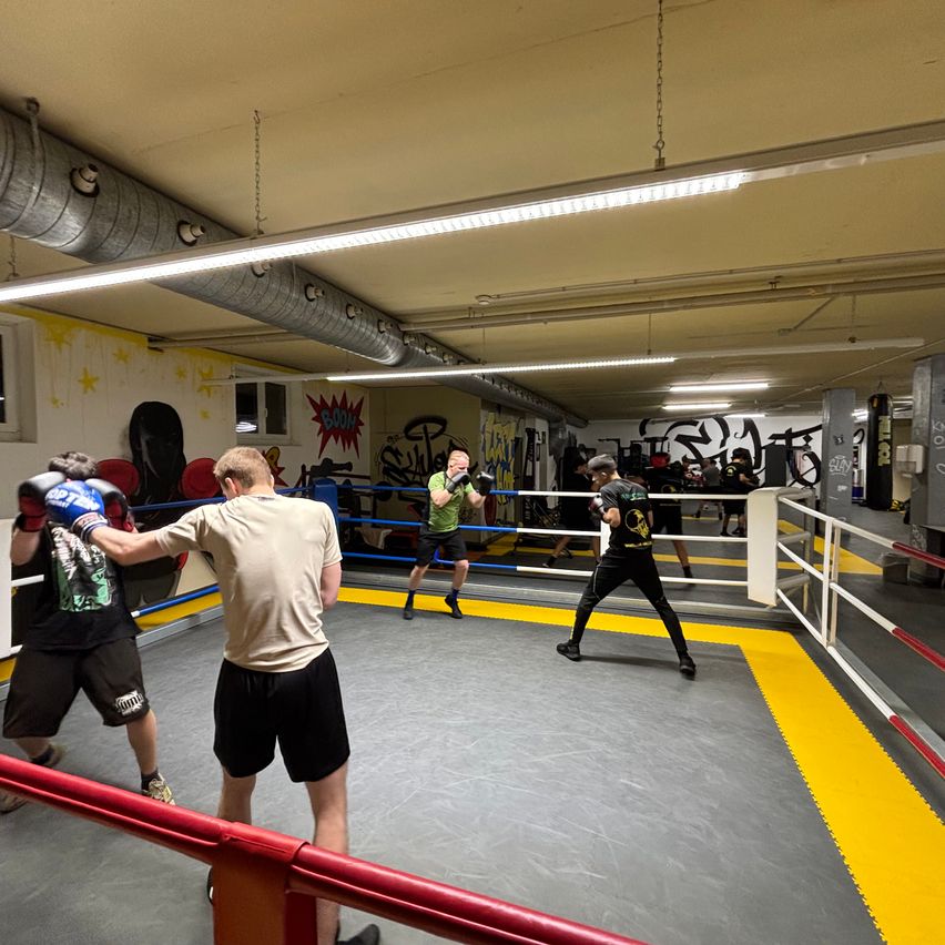 Mehrere Männer üben Boxen in einem Ring mit gelben und weißen Seilen. Zwei kämpfen aktiv, während andere stehen und zuschauen. Die Halle hat Graffiti an den Wänden.