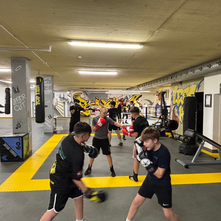 Eine Gruppe junger Männer übt Boxen in einem Fitnessstudio mit Graffiti an den Wänden, gelben Bodenmarkierungen und verschiedenen Boxausrüstungen.