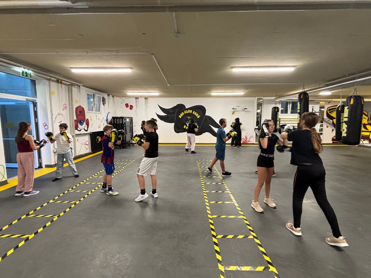 In einem Fitnessstudio üben mehrere Kinder Boxen mit Handschuhen und Schutzausrüstung. Die Halle hat eine Wandmalerei.