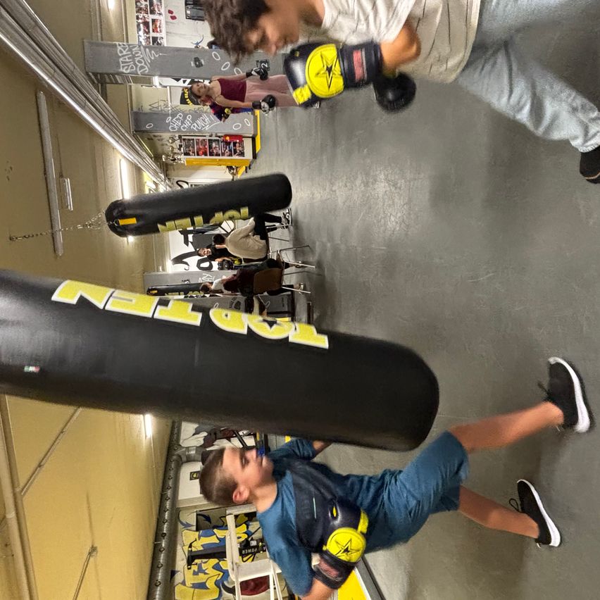 Zwei junge Männer in einem Fitnessstudio, einer übt Boxen mit einem Schlagsack, der andere beobachtet. Sie tragen beide Boxhandschuhe.