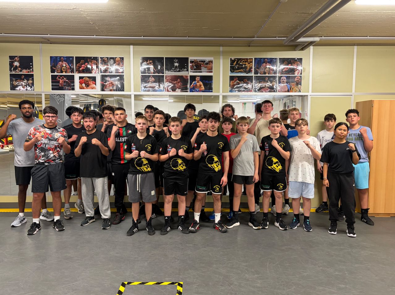 Eine Gruppe junger Männer in schwarzen und grauen Outfits posiert für ein Foto in einem Fitnessstudio. Sie haben gelbe Totenkopf-Designs auf ihren Kleidungsstücken. Sie stehen in einer Reihe, wobei einige von ihnen ihre Fäuste zeigen. Dahinter befindet sich eine Glaswand mit einem Spiegel und Bildern von Boxern darauf.