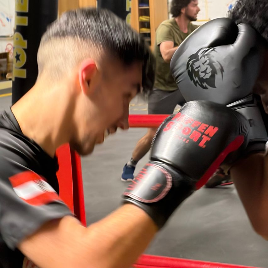Zwei Männer in einem Boxstudio, einer trägt schwarze Boxhandschuhe und der andere ein grünes Shirt. Ein anderer Mann beobachtet hinter den Seilen.