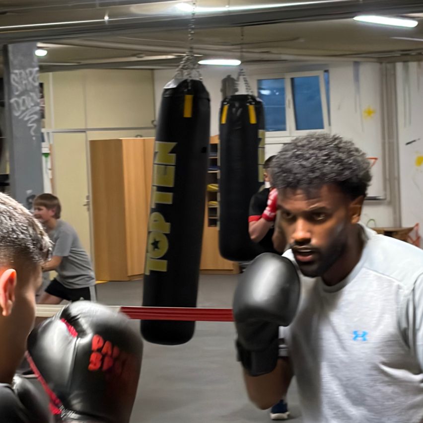 Zwei Männer in einem Fitnessstudio mit Boxhandschuhen. Ein Mann schaut den anderen an, der sich darauf vorbereitet, einen Boxsack zu schlagen.