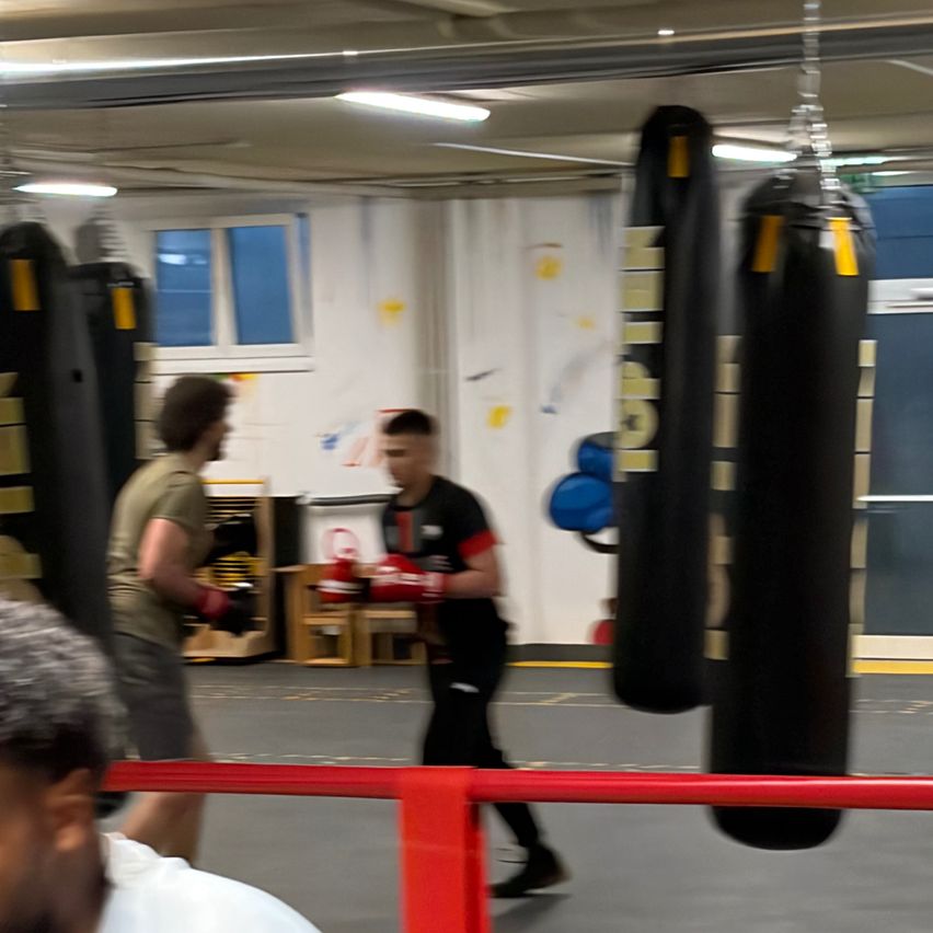 Zwei Männer in einem Fitnessstudio mit Boxhandschuhen. Einer schlägt auf einen hängenden Beutel, während ein anderer zusieht.