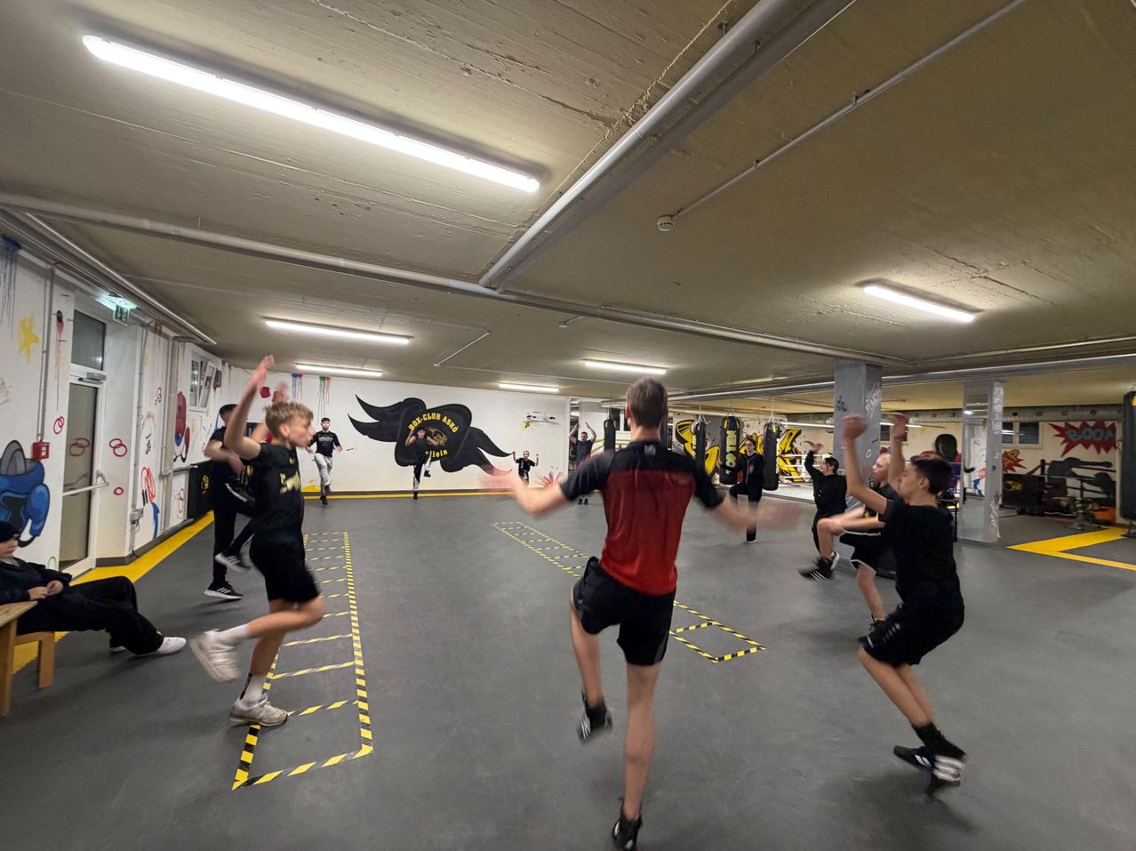 Eine Gruppe von Menschen in einem Fitnessstudio mit einem gelben Boden, die an einem Trainingsprogramm teilnehmen. Sie tragen Turnschuhe und schwarze T-Shirts. An der Wand befindet sich eine Wandmalerei.
