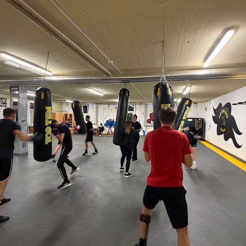 In einem Fitnessstudio üben mehrere Personen Boxen. Sie schlagen an Boxsäcken, die von der Decke hängen. Das Fitnessstudio hat gelbe Linien auf dem Boden und Graffiti an den Wänden.