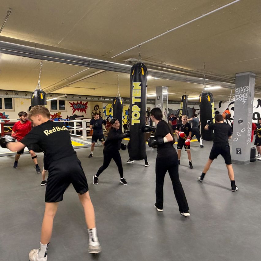 Eine Gruppe von Menschen boxt in einem Fitnessstudio. Sie tragen Boxhandschuhe und Uniformen. Einige schlagen auf Boxsäcke. Das Fitnessstudio hat weiße Wände und Graffiti.