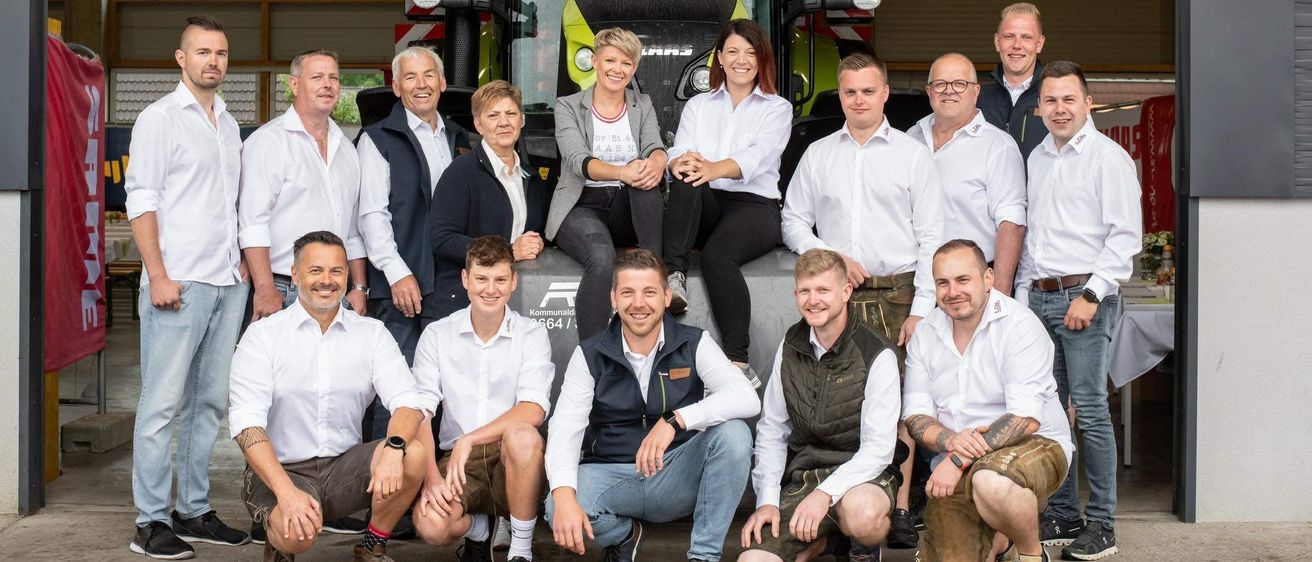 Eine Gruppe von Menschen, darunter Männer und Frauen, posiert für ein Foto vor einem Traktor. Sie tragen alle weiße Hemden und einige haben Brillen. Ein Banner zeigt 'LTH Landtechnik Huttner'.