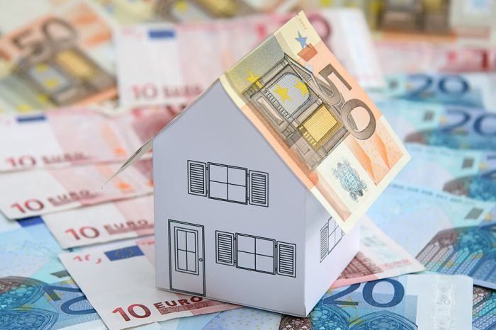 Ein kleines Papierhaus sitzt auf einem Haufen 10 und 20 Euro Scheinen und symbolisiert finanzielle Stabilität und Immobilieninvestitionen.