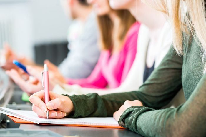 Ein Klassenzimmer, in dem eine Frau in ein Notizbuch mit einem Stift schreibt, während andere im Hintergrund verschwommen sind.