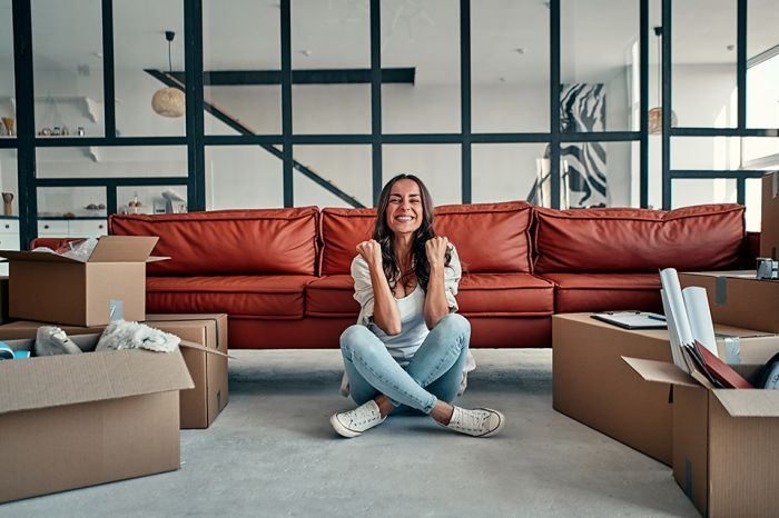 Eine Frau sitzt vor einer roten Ledercouch auf dem Boden, umgeben von Umzugskartons. Sie lächelt und scheint für ein Foto zu posieren.