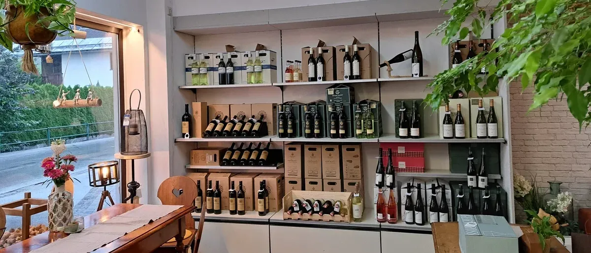 Ein Raum mit Regalen voller Weinflaschen. Es gibt einen Holztisch und Stühle. Auf der rechten Seite des Raumes steht eine Pflanze.