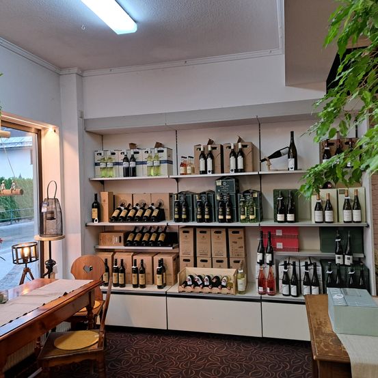 Ein Weinladen mit einem hölzernen Tisch und Stühlen vor einem großen Fenster, mit Regalen voller Weinflaschen und Kisten, einer Deckenleuchte und einer grünen Pflanze.