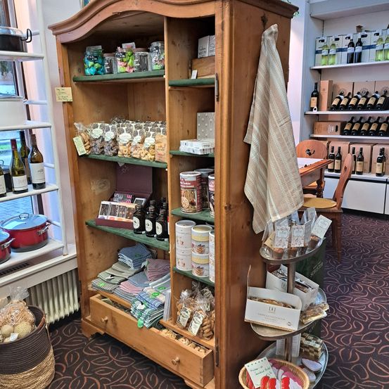 Ein hölzerner Schrank voller Snacks, Gläser und Tüten auf Regalen. Ein Handtuch hängt am Schrank. Ein Tisch und Stühle sind im Hintergrund. Flaschen Wein stehen auf Regalen. Ein Korb mit Gegenständen steht auf dem Boden.